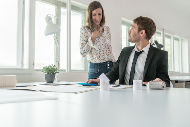 Jonge zakenman zit aan zijn bureau en bekijkt financieel verslag en zijn vrouwelijke partner staat naast hem in een discussie.