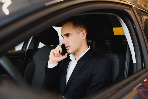 Jonge zakenman zit in luxeauto en praat over de telefoon.