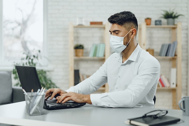 Jonge zakenman zittend aan zijn bureau met medische masker op, portret