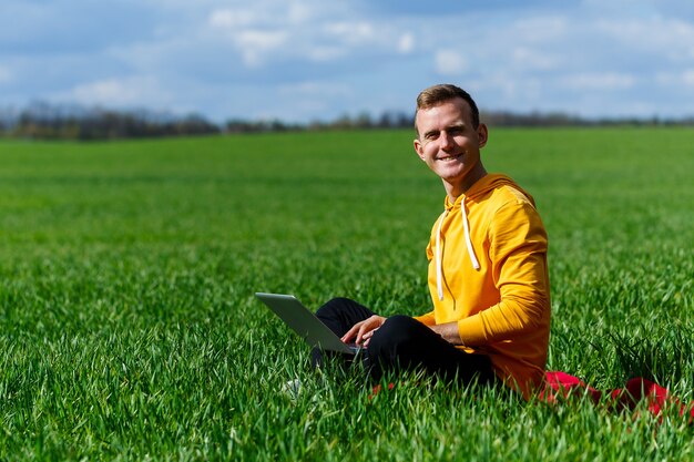 Jonge zakenman zittend op groen gras en met behulp van laptopcomputer. Knappe man aan het werk met computer in park op zonnige zomerdag. Buitenshuis natuur reis en ontspanning. Freelance werkconcept