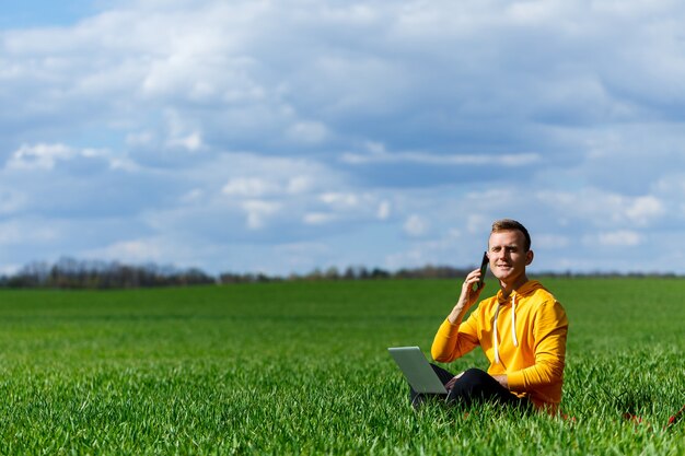 Jonge zakenman zittend op groen gras praten aan de telefoon en het gebruik van een laptopcomputer. knappe man aan het werk met een computer in een park op een zonnige zomerdag. freelance werkconcept