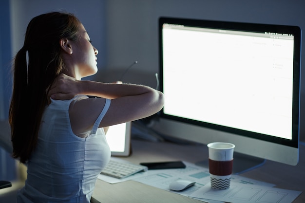 Jonge zakenvrouw die een stijve nek kneedt terwijl ze op een gloeiend computerscherm zit in een donker kantoor