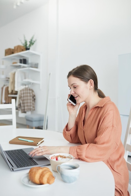 Jonge zakenvrouw die laptop gebruikt tijdens haar gesprek op mobiele telefoon, terwijl ze aan de tafel in de keuken werkt