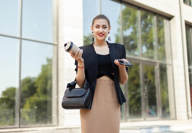 Jonge zakenvrouw die telefoon vasthoudt en onderweg koffie drinkt