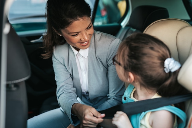 Jonge zakenvrouw helpt haar dochter om veiligheidsgordels in de auto vast te maken terwijl het meisje op een veiligheidskinderzitje zit.