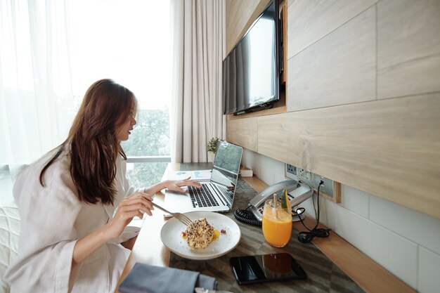 Jonge zakenvrouw in badjas ontbijten en werken op laptop aan tafel in hotelkamer