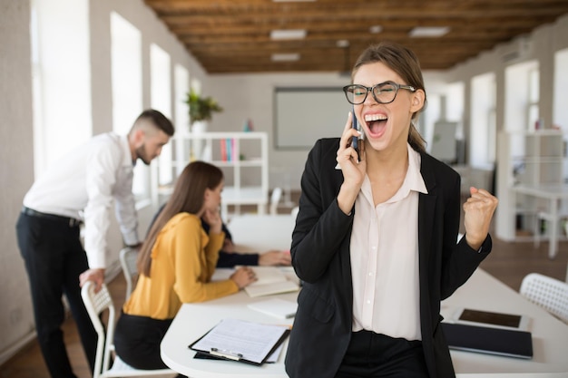 Jonge zakenvrouw in bril en shirt die emotioneel schreeuwt terwijl ze op mobiel op kantoor praat met collega's op de achtergrond