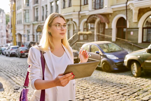 Jonge zakenvrouw in de stad met behulp van digitale tablet. Mooie lachende vrouw in witte jas met bril kijken naar tablet-monitor, stedelijke stijl achtergrond