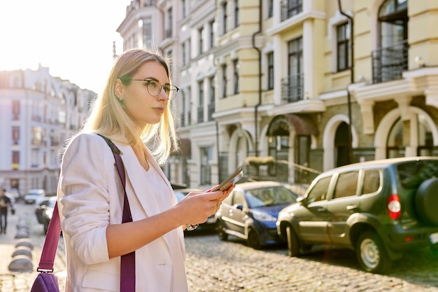 Jonge zakenvrouw in de stad met behulp van digitale tablet. Mooie lachende vrouw in witte jas met bril kijken naar tabletmonitor, kopieer ruimte, stedelijke stijl achtergrond
