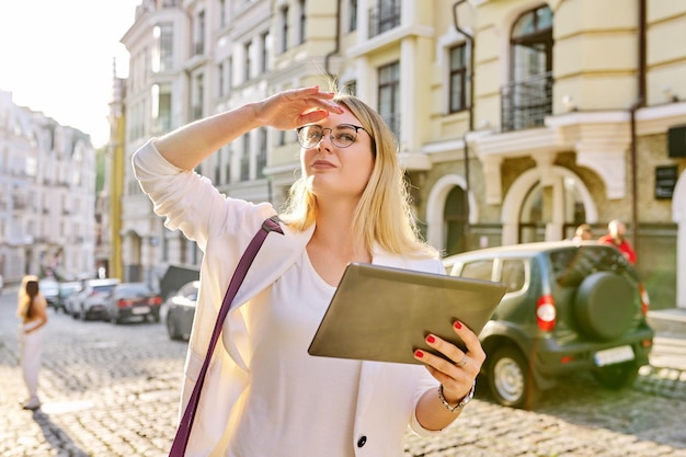 Jonge zakenvrouw in de stad met behulp van digitale tablet Mooie lachende vrouw in witte jas met bril op zoek in de verte stedelijke stijl achtergrond