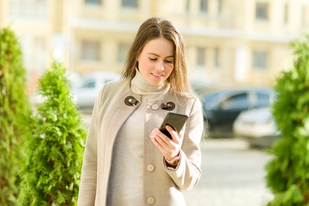 Jonge zakenvrouw in herfst stad in jas met smartphone op straat. Stadsportret van succesvolle modieuze mooie vrouw