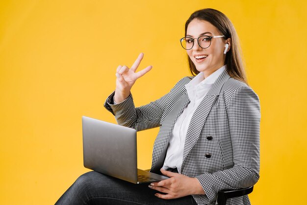 Jonge zakenvrouw in pak met een bril die zit en een laptop op gele achtergrond gebruikt