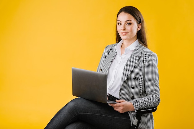 Jonge zakenvrouw in pak met een bril die zit en een laptop op gele achtergrond gebruikt