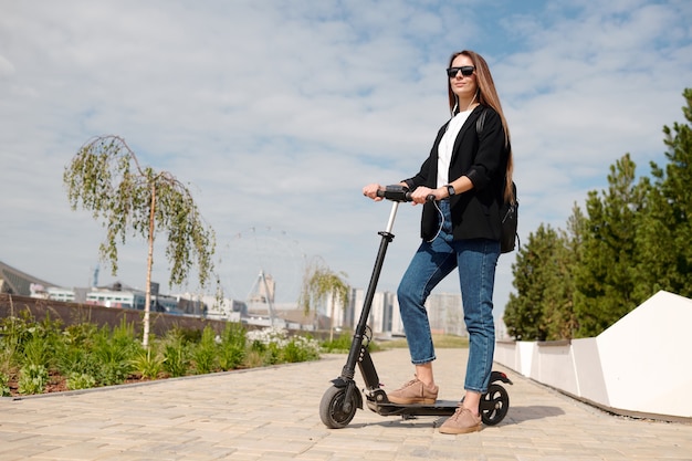 Jonge zakenvrouw in vrijetijdskleding en zonnebril die op een elektrische scooter staat in een stedelijke omgeving tegen huizen, bomen en bewolkte hemel