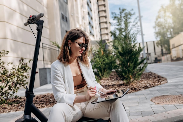 Jonge zakenvrouw in wit pak zittend op een elektrische scooter en bezig met digitale tablet in de stad met moderne architectuur