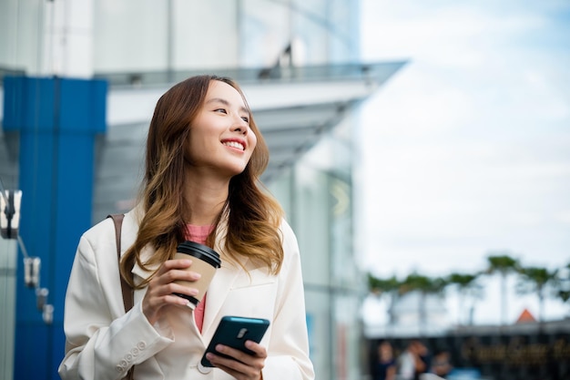 Jonge zakenvrouw lachend met mobiele telefoon met koffie meenemen om vroeg in de ochtend naar het werk te gaan, Aziatische zakenvrouw met smartphone en kopje koffie staande tegen straatgebouw in de buurt van kantoor