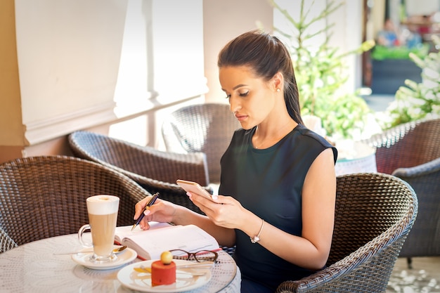 Jonge zakenvrouw maakt aantekeningen in notitieblok van smartphone kijken naar koffiepauze in café werken