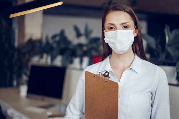 Jonge zakenvrouw met gezichtsmasker die op kantoor staat en klembord met documenten vasthoudt