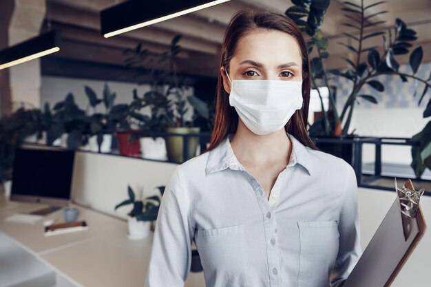 Jonge zakenvrouw met gezichtsmasker die op kantoor staat en klembord met documenten vasthoudt