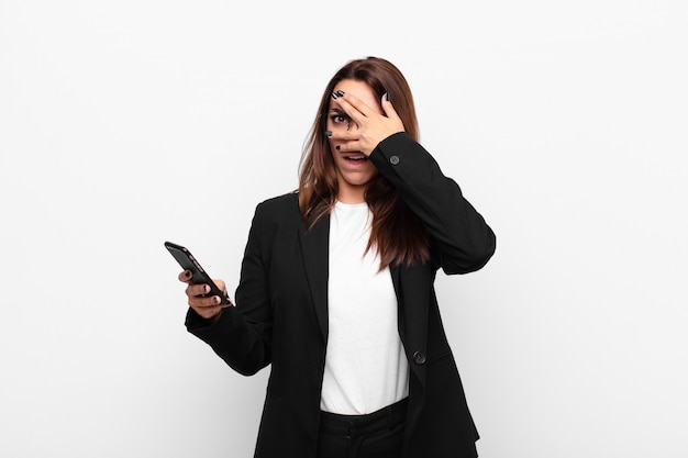 Jonge zakenvrouw op zoek geschokt, bang of doodsbang, die gezicht bedekt met hand en gluren tussen vingers en een mobiele telefoon te houden