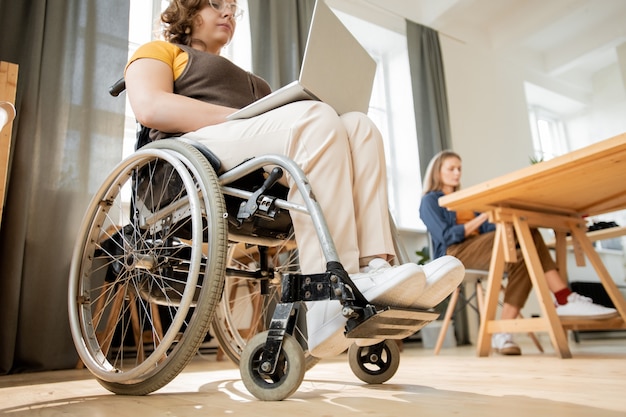 Jonge zakenvrouw uitschakelen met laptop op knieën zittend in een rolstoel op kantoor en surfen in het net tegen haar collega die aan tafel werkt