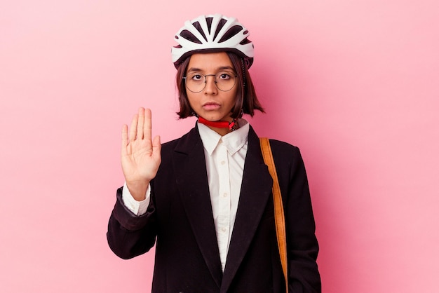 Jonge zakenvrouw van gemengd ras die een fietshelm draagt, geïsoleerd op een roze achtergrond die staat met uitgestrekte hand die een stopbord toont, waardoor je wordt verhinderd.