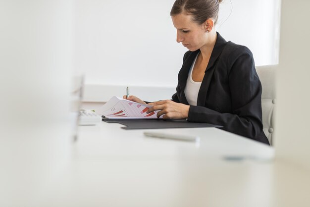 Jonge zakenvrouw zit aan haar heldere bureau en werkt aan papierwerk en financieel rapport vertegenwoordigd in grafieken en diagrammen.