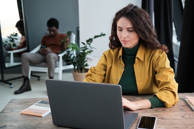 Jonge zakenvrouw zittend aan tafel en werkend op laptop met man zittend op de achtergrond op kantoor