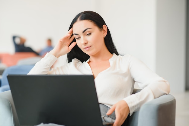 Jonge zakenvrouw zittend op de bank en werkend met laptop in de wachtzaal van het zakencentrum