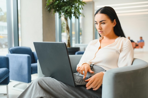 Jonge zakenvrouw zittend op de bank en werkend met laptop in de wachtzaal van het zakencentrum