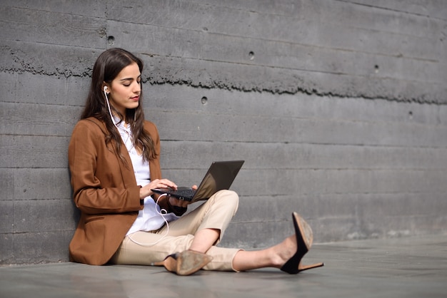 Jonge zakenvrouw zittend op de vloer op zoek naar haar laptop computer. mooie vrouw met formele slijtage met behulp van koptelefoon.