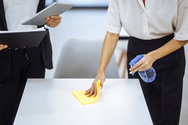 Jonge zakenvrouwen maken de werkplek schoon, veeg het bureau af met een gele doek. Collega's desinfecteren het werkoppervlak met een ontsmettingsspray om de verspreiding van covid-19 te stoppen.