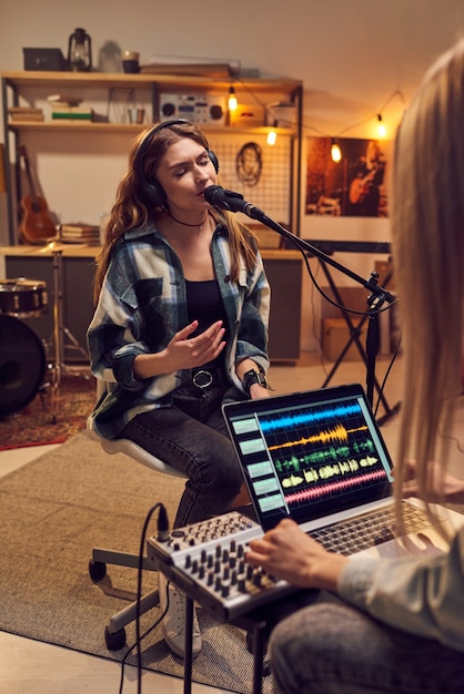 Jonge zangeres in koptelefoon zingt in microfoon terwijl vrouw laptop gebruikt om haar lied op te nemen in de studio