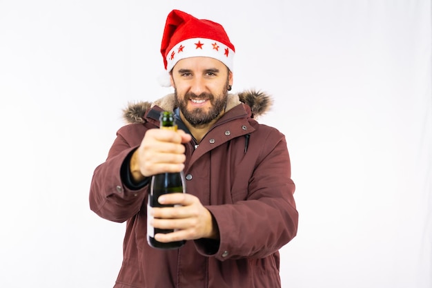 Jonge, zeer gelukkige blanke man met rode kerstmuts die een fles champagne opent op een witte achtergrond, gekleed in een winterjas