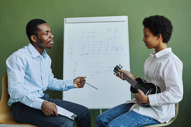 Jonge zelfverzekerde afro-amerikaanse leraar die vragen beantwoordt van schoolmeisje tijdens les met ukelele wh