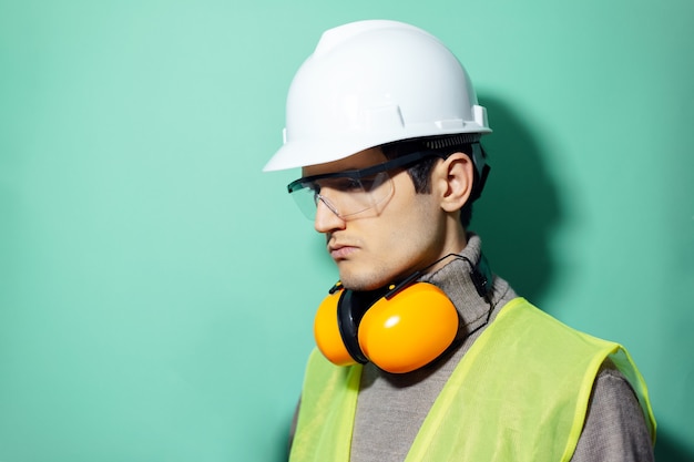 Jonge zelfverzekerde bouwersman, ingenieur, bouwveiligheidskoptelefoon op nek, harde helm, veiligheidsbril en reflecterend jasje op muur van aqua menthe-kleur.