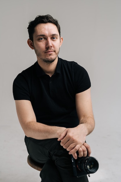 Jonge zelfverzekerde man fotograaf in zwart t-shirt met digitale camera, zittend op een stoel. Studiofoto op geïsoleerde witte achtergrond.