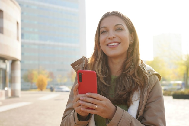 Jonge zelfverzekerde vrouw die smartphone over de achtergrond van de stad gebruikt, lacht en kijkt naar de camera