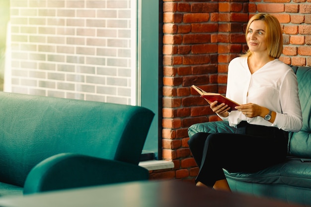 Jonge zelfverzekerde vrouw ondernemer geniet van de werkpauze zittend op een leren bank in kantoor interieur, intelligente geweldige vrouwelijke advocaat in formele slijtage een boek lezen.