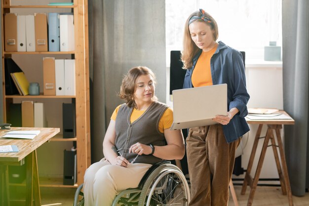 Jonge zelfverzekerde zakenvrouw met laptop die informatie toont voor een rapport of zakelijk project aan haar invalide collega in rolstoel