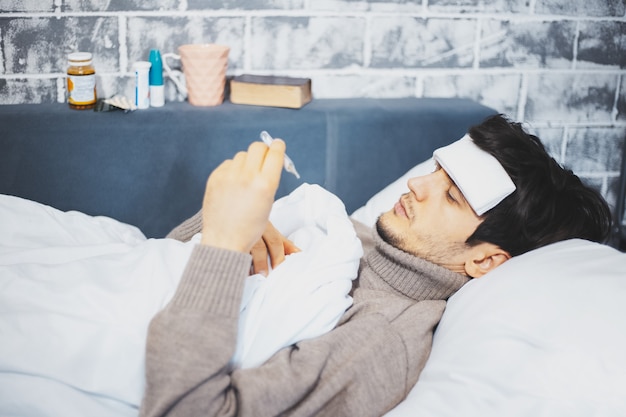 jonge zieke man liggend op bed met thermometer in de hand op muur van andere medicijnen.