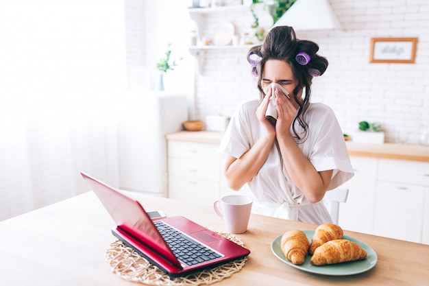 Jonge zieke vrouw houdt weefsel rond neus. Kijkend op laptop. Zieke huishoudster die film kijkt. Influenza. Alleen thuis. Ochtend daglicht.