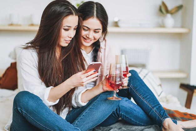 Jonge zorgeloze brunette meisjes vrienden in casual met glazen wijn met behulp van mobiele en plezier samen op home party