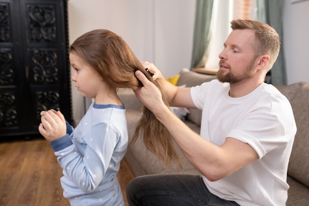 Jonge, zorgvuldige vader met haarborstel lang haar van zijn schattige dochtertje borstelen zittend op de bank voor haar in de woonkamer
