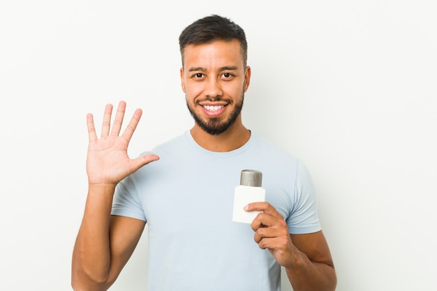 Jonge Zuid-Aziatische mens die een aftershavecrème houdt die vrolijk tonend nummer vijf met vingers glimlacht.