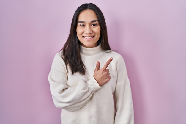 Jonge zuid-aziatische vrouw die vrolijk over een roze achtergrond staat met een glimlach op het gezicht wijzend met hand en vinger naar de zijkant met een gelukkige en natuurlijke uitdrukking