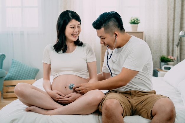 Jonge zwangere Aziatische Japanse vrouw en knappe man zittend op bed. man toekomstige vader met behulp van stethoscoop luister naar vrouw blote buik met ongeboren baby erin. gelukkige vaders dag beste cadeau ooit.