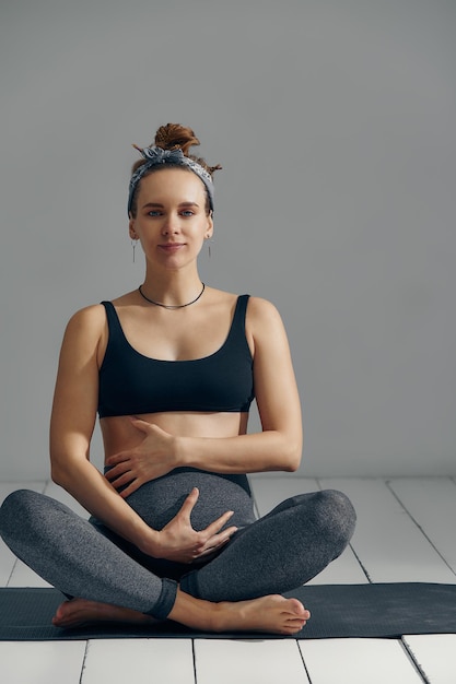 Jonge zwangere duizendjarige vrouw die op de mat zit, raakt haar buik aan na het uitvoeren van prenatale en meditatie-oefeningen tijdens een yogales concept van leven en moederschap