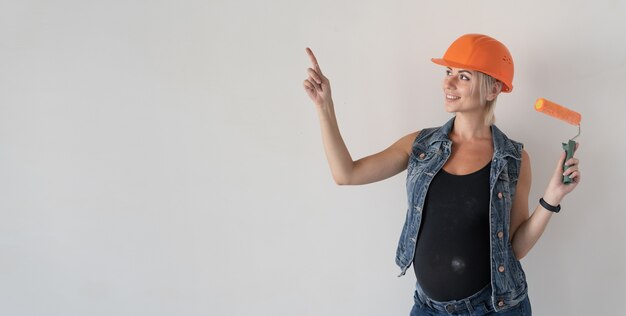 Jonge zwangere mooie vrouwenbouwer in een oranje beschermende helm op haar hoofd. Zwanger meisje. een verfroller vasthouden en met een vinger omhoog wijzen naar een lege ruimte voor tekst