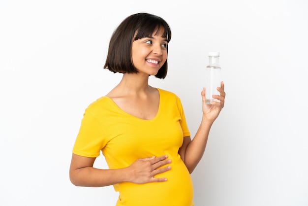 Jonge zwangere vrouw die een fles water houdt die op witte achtergrond wordt geïsoleerd en naar de zijkant kijkt